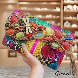May include: A colorful clutch with a snakeskin pattern in shades of pink, yellow, green, and blue. The flap is adorned with decorative stones in various colors and a gold dragonfly. The clutch has a gold tassel and the text "GonolD".