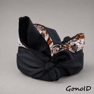 May include: A black fabric head covering featuring a knotted design and a contrasting interior lining with a white and brown floral pattern. The word GonolD is printed in black in the bottom right corner of the image.