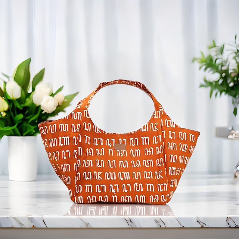 Peut inclure: Un sac fourre-tout orange avec un motif blanc et une poign&eacute;e circulaire. Le sac a une &eacute;tiquette en cuir marron. Le sac est pos&eacute; sur une surface en marbre blanc.