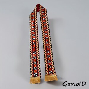 Puede incluir: Una larga bufanda decorativa con un fondo rojo y intrincados patrones bordados en negro, blanco y naranja. La bufanda tiene flecos dorados en los extremos. La palabra "GonolD" está impresa en la esquina inferior derecha.