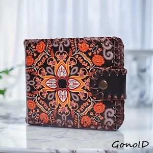 May include: A square, black wallet with an intricate floral pattern in orange, yellow, and white. The wallet has a leather strap closure with a metal snap. The edges are stitched with brown thread. The word "GonolD" is printed in the bottom right corner.