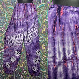 May include: Purple tie-dye pants with a relaxed fit. The pants feature a mix of dark and light purple hues with white stripes and floral accents. The waistband is elasticized. The fabric appears lightweight and suitable for casual wear.