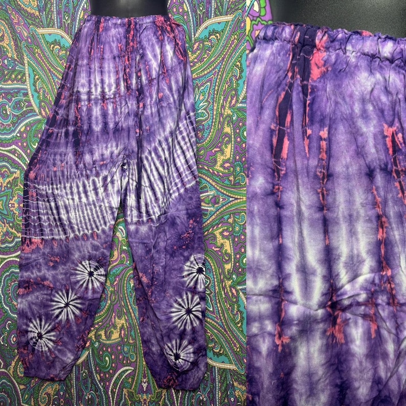 May include: Purple tie-dye pants with a relaxed fit. The pants feature a mix of dark and light purple hues with white stripes and floral accents. The waistband is elasticized. The fabric appears lightweight and suitable for casual wear.
