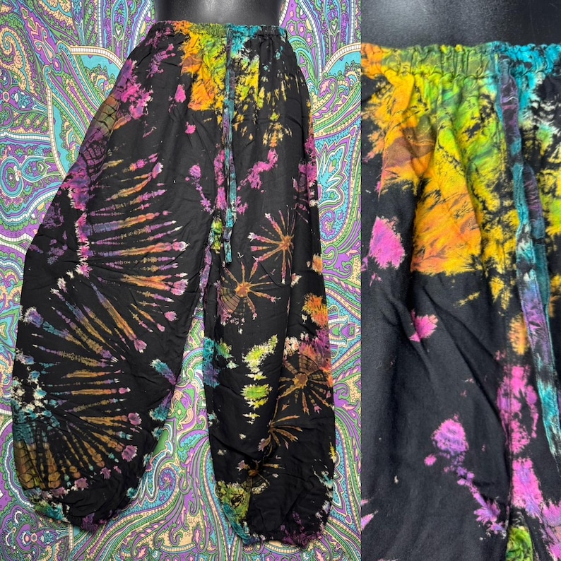 May include: Black tie-dye pants with a colorful burst pattern in shades of pink, orange, yellow, green, and purple. The pants have an elastic waistband and a drawstring. The fabric appears to be lightweight and flowing.