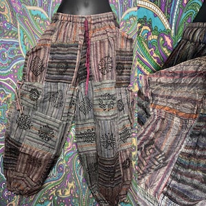 Puede incluir: Pantalones patchwork en tonos grises, marrones y morados, con una mezcla de paneles de tela a rayas y estampados. Los pantalones tienen cintura con cordón y bolsillos, con diseños florales y geométricos. El estilo es bohemio.