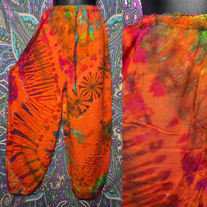 May include: Orange tie-dye pants with a drawstring waist. The pants feature a vibrant pattern of orange, pink, and green hues. The design includes radial and floral-like patterns. The pants are made of a lightweight fabric.
