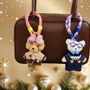 Puede incluir: Dos llaveros de osos de peluche. Uno es marrón claro con un lazo rosa, un suéter y una cámara. El otro es blanco con gafas y un peto. Ambos tienen correas de cuerda coloridas y cuelgan frente a un bolso marrón.