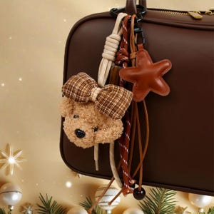 May include: A brown teddy bear keychain with a plaid bow, a brown leather star, and various cords and straps. The keychain is attached to a brown leather bag with a black clasp. The background is a soft beige with decorative elements.