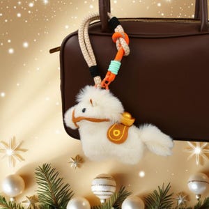 Pode incluir: Um chaveiro de cavalo de pelúcia branco com sela marrom e detalhes dourados. O chaveiro está preso a uma bolsa de couro marrom com uma corda bege e laranja. Decorações de Natal no fundo.