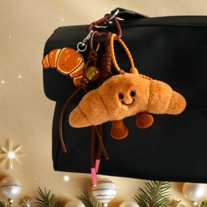 Puede incluir: Un llavero de croissant de peluche marrón con una cara sonriente y pies pequeños, colgado de un bolso negro. El llavero también incluye un dije de croissant de dibujos animados y cuentas decorativas. El fondo es de un color cálido.