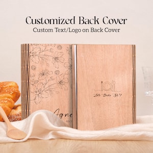 K&ouml;nnte beinhalten: Ein h&ouml;lzernes Rezeptbuch mit floralem Design und dem Text "Customized Back Cover". Das Buch ist ge&ouml;ffnet und zeigt die Innenseiten. Auf einer Seite steht "Agne RECI" und auf der anderen Seite "Lets Bake Sh!t".