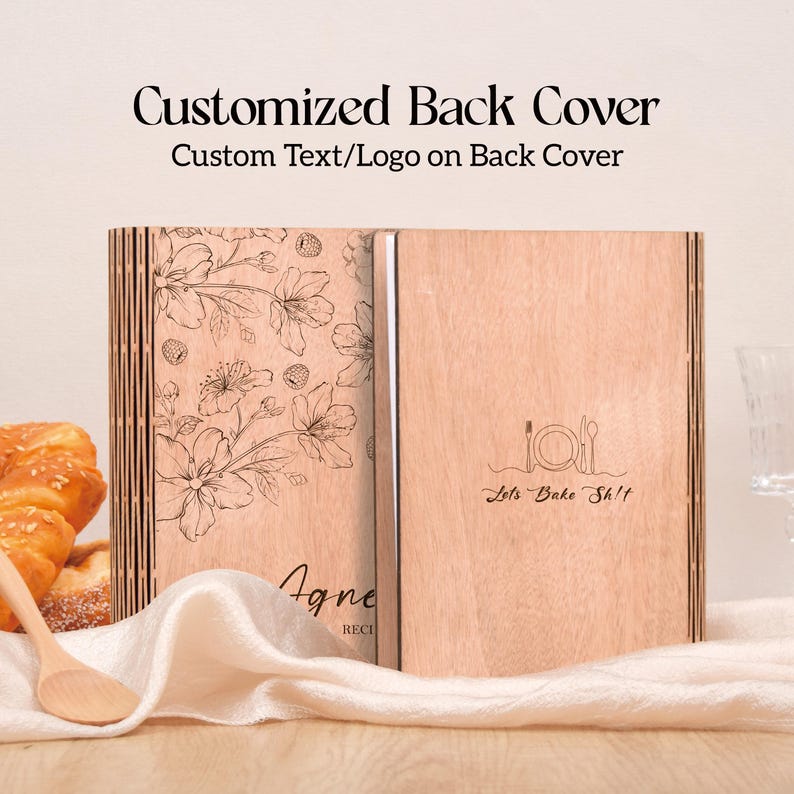 K&ouml;nnte beinhalten: Ein h&ouml;lzernes Rezeptbuch mit floralem Design und dem Text "Customized Back Cover". Das Buch ist ge&ouml;ffnet und zeigt die Innenseiten. Auf einer Seite steht "Agne RECI" und auf der anderen Seite "Lets Bake Sh!t".