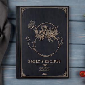 Könnte beinhalten: Ein schwarzes, rechteckiges Rezeptbuch mit goldener Linienzeichnung einer Teekanne voller Blumen und einem Schmetterling. Der Text lautet EMILY'S RECIPES, Made with love, Shared with joy. Das Design enthält dekorative Verzierungen.
