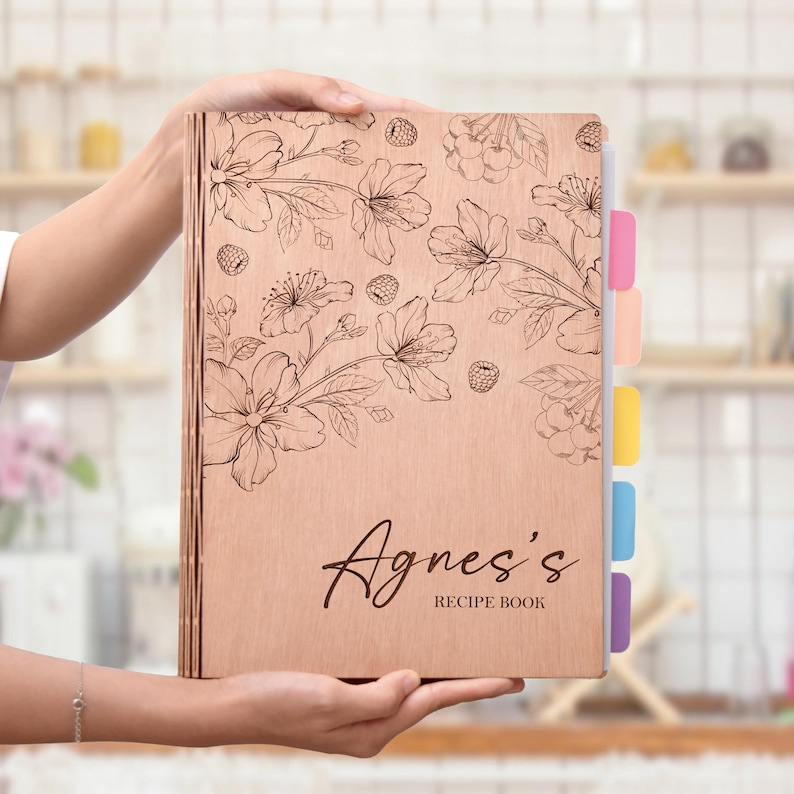 K&ouml;nnte beinhalten: Ein h&ouml;lzernes Rezeptbuch mit floralem Design und dem eingravierten Namen "Agnes's" auf dem Cover. Das Buch hat farbige Registerkarten und wird offen gehalten. Das florale Design umfasst Blumen und Beeren.