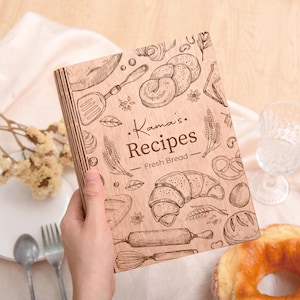 May include: A wooden recipe book with the text "Kama's Recipes - Fresh Bread" and illustrations of baked goods. The cover features a croissant, rolling pin, and other bread-related designs. The book is held in a person's hand.