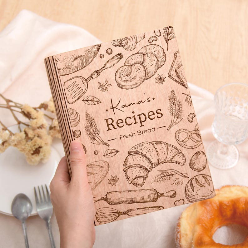 K&ouml;nnte beinhalten: Ein h&ouml;lzernes Rezeptbuch mit dem Text "Kama's Recipes - Fresh Bread" und Illustrationen von Backwaren. Das Cover zeigt ein Croissant, eine Teigrolle und andere brotbezogene Designs. Das Buch wird in der Hand einer Person gehalten.