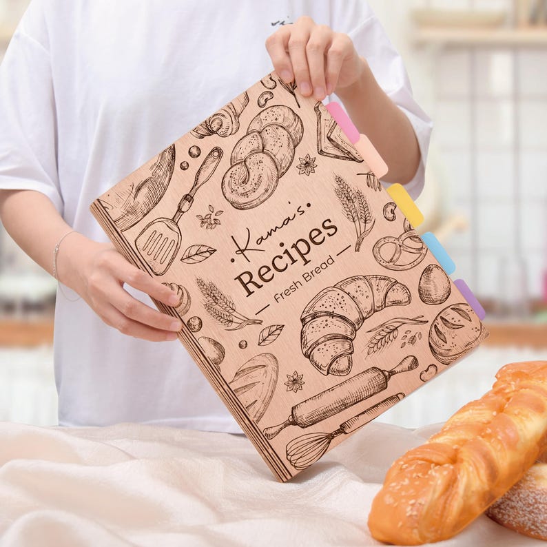 K&ouml;nnte beinhalten: Ein Rezeptordner aus Holz mit dem Text "Kama's Recipes - Fresh Bread" und Illustrationen von Backwaren. Der Ordner hat farbige Registerkarten und wird von einer Person gehalten.