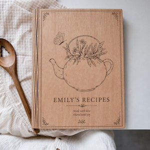 Puede incluir: Un libro de recetas rectangular de madera con acabado marrón claro. La portada presenta una ilustración grabada con láser de una tetera llena de flores y una mariposa. El texto dice: EMILY'S RECIPES, Made with love, Shared with joy. Una cuchara de madera está al lado.