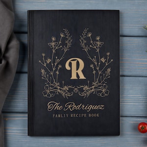 May include: A black rectangular recipe book with a gold-coloured letter R centred within a floral wreath design. Below the wreath, the text reads The Rodriguez FAMLIY RECIPE BOOK in gold lettering. The cover features a vertical lined spine detail.