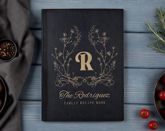 May include: A black rectangular recipe book with a gold-colored letter R centered within a floral wreath design. Below the wreath, the text reads The Rodriguez FAMLIY RECIPE BOOK in gold lettering. The cover features a vertical lined spine detail.