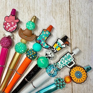 May include: A collection of decorative pens with various toppers, including a pink cowboy hat, a brown cowboy hat, a turquoise boot, and a coffee cup. The pens have colorful beads and accents, with a wooden background.