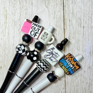 May include: A collection of black and white pens with decorative tops. The tops feature various designs and phrases, including "Fresh Out of Fucks" and "But did you Die?" The pens have a mix of patterns and colors.