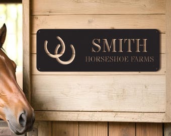 May include: Black rectangular metal sign with the name "SMITH" and "HORSESHOE FARMS" in gold lettering. Two interlocking horseshoe cutouts are on the left. The sign is mounted on a wooden wall, with a horse's head visible on the left.