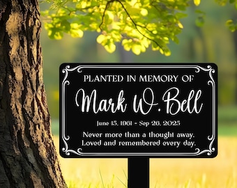 Puede incluir: Un letrero conmemorativo negro con texto blanco, plantado en una zona de césped. El letrero dice "Plantado en memoria de Mark W. Bell" con las fechas 15 de junio de 1961 - 26 de septiembre de 2025, y un mensaje cariñoso.