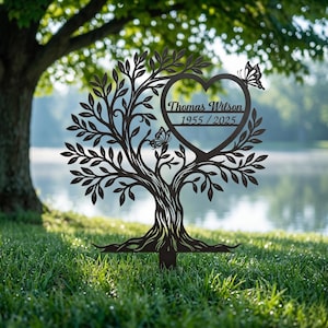 May include: A dark metal tree sculpture with a heart-shaped cutout and butterfly accents. The heart contains the inscription "Thomas Wilson 1955 / 2025". The tree is set in a grassy area with a blurred background of trees and water.