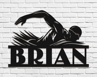 May include: Black metal wall art depicting a swimmer in the freestyle stroke. The swimmer is wearing goggles and a cap. The name "BRIAN" is displayed in bold letters below the swimmer. The art is mounted on a white brick wall.