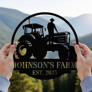 Peut inclure: Panneau de ferme en métal noir représentant un tracteur et une personne. Le panneau est circulaire et porte l'inscription "JOHNSON'S FARM" et "EST. 2025". L'arrière-plan montre un paysage de montagne flou.