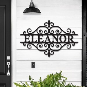 Può includere: Cartello in metallo nero con il nome "ELEANOR" in un carattere decorativo. Il cartello è montato su una parete di legno bianca e presenta volute ornate. Sopra il cartello è appesa una lampada nera.