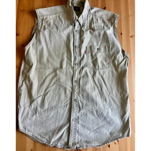 May include: A sleeveless, button-down shirt in a light tan color. The shirt has a collared neckline, a single chest pocket, and a red logo tag. The shirt is made of a durable fabric and has a classic, casual style.