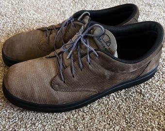 May include: A pair of brown suede casual shoes with black soles and laces. The shoes have a low-profile design and the brand name "ellesse" is visible on the tongue.
