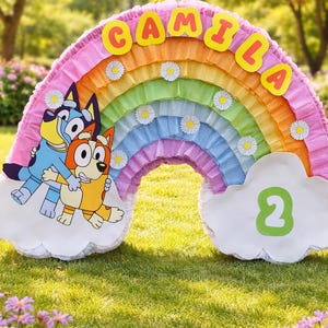May include: A colorful rainbow piñata with the name "Camila" in yellow letters. The piñata features cartoon characters and a cloud with the number "2". Daisies decorate the rainbow, set on a grassy lawn.