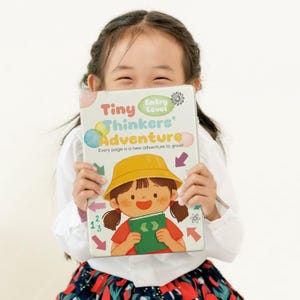 Puede incluir: Un niño sostiene un libro titulado "Tiny Thinkers' Adventure". La portada del libro presenta una ilustración de dibujos animados de un niño con un sombrero amarillo y sosteniendo un libro verde. La portada también incluye gráficos coloridos y el texto "Every page is a new adventure to grow!"