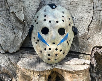 May include: A weathered, off-white hockey mask with black straps and blue chevron markings. The mask has numerous small, evenly spaced holes and is resting on a textured, wooden surface. A prop for Halloween or cosplay.