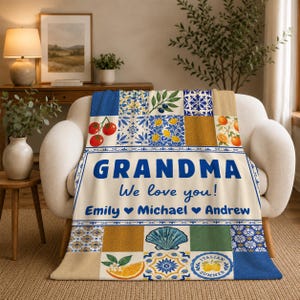 May include: A cream-colored throw blanket featuring a Mediterranean-inspired tile pattern with lemons, tomatoes, oranges, and seashells. The text reads GRANDMA We love you! Emily, Michael, and Andrew, separated by blue heart icons in a bold font.