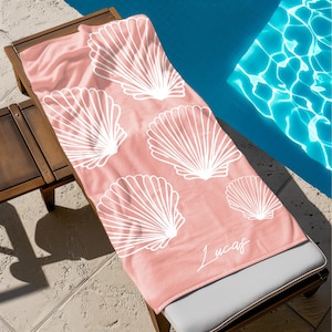 May include: A rectangular beach towel in a soft peach color featuring a repeating pattern of white line-art seashells. The name Lucas is printed in a white cursive script font in the bottom right corner of the fabric surface.