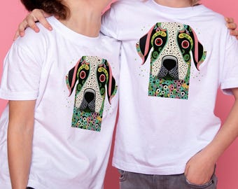 May include: Two white t-shirts with a colorful dog graphic. The dog design features a white face with black spots, red eyes, and green, pink, and black accents. The dog's neck is surrounded by a floral pattern.