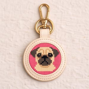May include: A round keychain featuring a cross-stitched pug dog design on a pink background. The keychain has a cream-colored leather border and a gold-colored clasp. The pug has a beige face, dark ears, and a black nose.