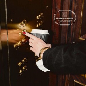 May include: A person's hand with red nail polish presses an elevator button, holding a black and white paper coffee cup. The person wears a gold bracelet and a black blazer with a white shirt cuff. The elevator panel is gold-toned.