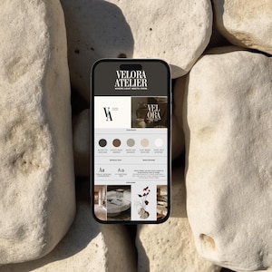 May include: A smartphone displaying a website design with the text "VELORA ATELIER" and a color palette including black, brown, and beige tones. The phone rests on a background of large, light-colored stones.