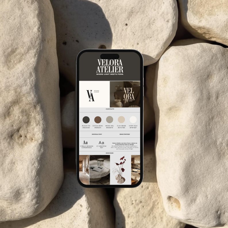 May include: A smartphone displaying a website design with the text "VELORA ATELIER" and a color palette including black, brown, and beige tones. The phone rests on a background of large, light-colored stones.