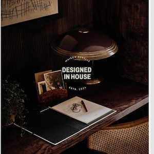 May include: A dark brown desk with a lamp, notepad, and stationery. The lamp has a brown shade and gold base. A notepad with a pen and a pair of glasses is on the desk. The text "MAISON AURELLE DESIGNED IN HOUSE ESTB. 2024" is visible.