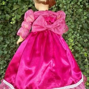 Puede incluir: Un vestido de sat&eacute;n rosa con una capa transparente y un lazo en la espalda. El vestido tiene un borde de encaje blanco y es perfecto para una mu&ntilde;eca.