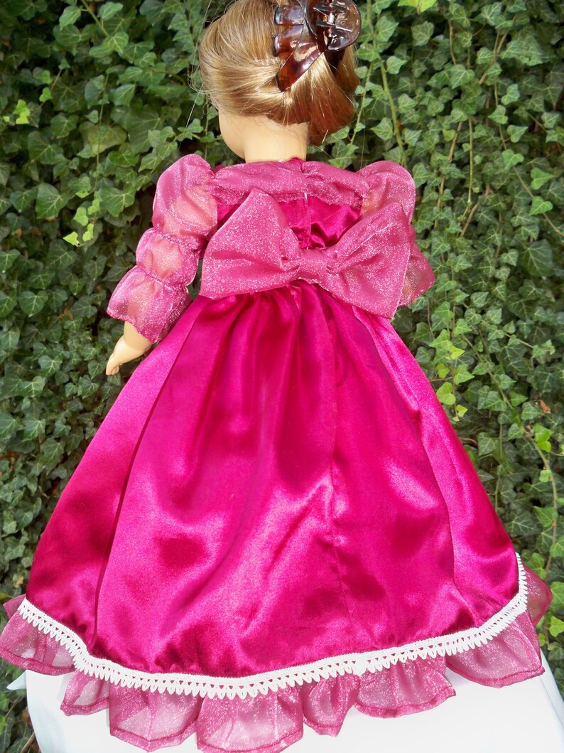 Puede incluir: Un vestido de sat&eacute;n rosa con una capa transparente y un lazo en la espalda. El vestido tiene un borde de encaje blanco y es perfecto para una mu&ntilde;eca.