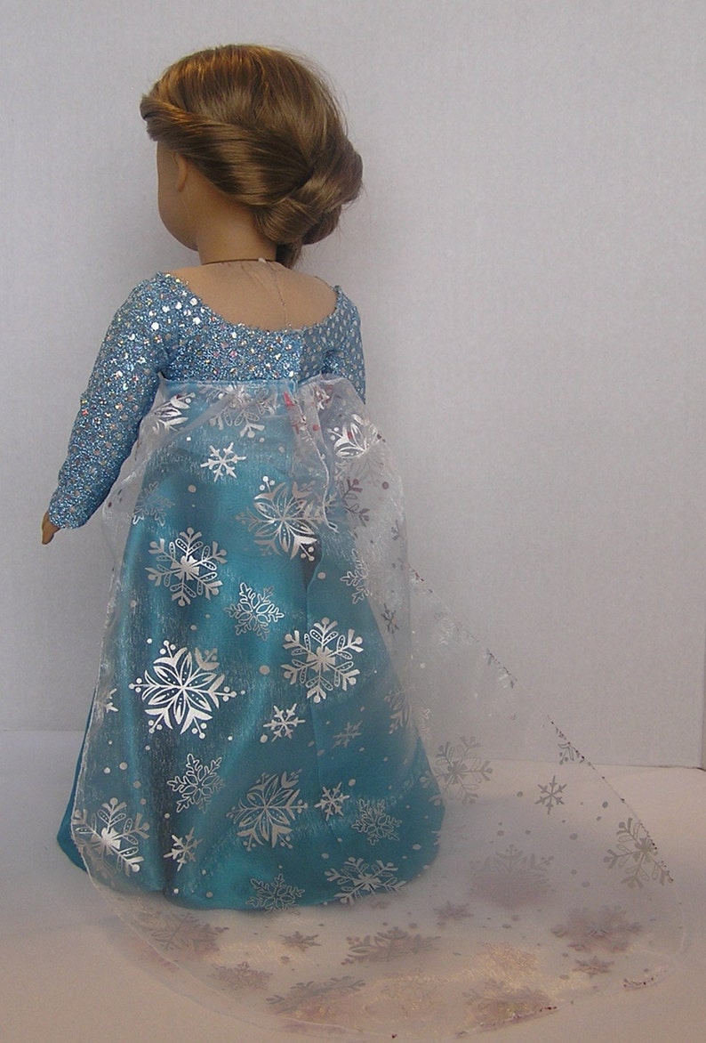 Puede incluir: Un vestido azul y plateado con estampado de copo de nieve y una superposici&oacute;n transparente. El vestido lo lleva una mu&ntilde;eca con el pelo casta&ntilde;o.