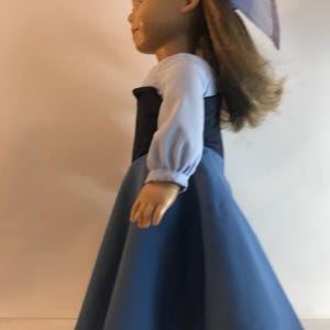 K&ouml;nnte beinhalten: Eine Puppe in einem blauen Kleid mit schwarzem Oberteil und langen &Auml;rmeln. Die Puppe hat lange, hellbraune Haare und eine blaue Schleife. Das Kleid hat einen weiten Rock und ist aus einem weichen Stoff gefertigt. Die Puppe tr&auml;gt schwarze Schuhe.