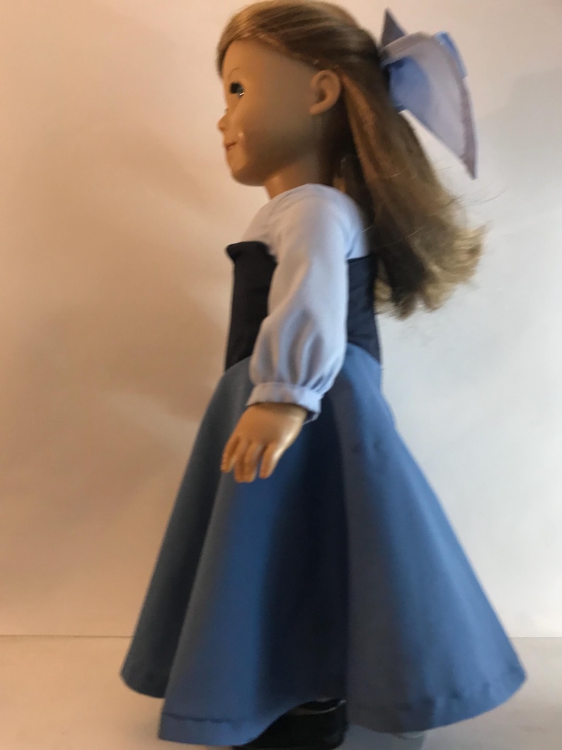 K&ouml;nnte beinhalten: Eine Puppe in einem blauen Kleid mit schwarzem Oberteil und langen &Auml;rmeln. Die Puppe hat lange, hellbraune Haare und eine blaue Schleife. Das Kleid hat einen weiten Rock und ist aus einem weichen Stoff gefertigt. Die Puppe tr&auml;gt schwarze Schuhe.